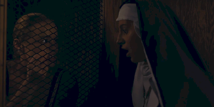 Woman with blonde hair looking through a wire mesh while a woman in a nun’s habit stands close on the other side, facing her in profile.
