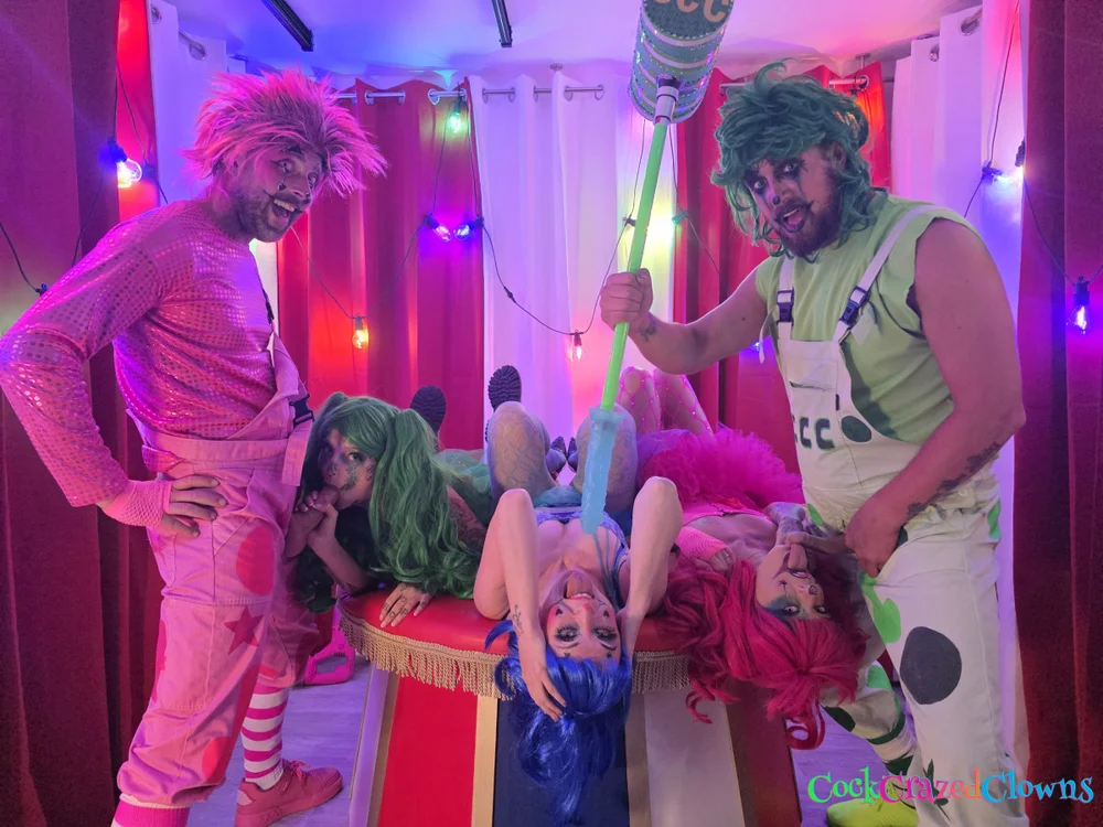 Woman with blue hair lying on her back on a red platform while two women in clown costumes perform oral sex on two men standing nearby; one man holds a large prop mallet.