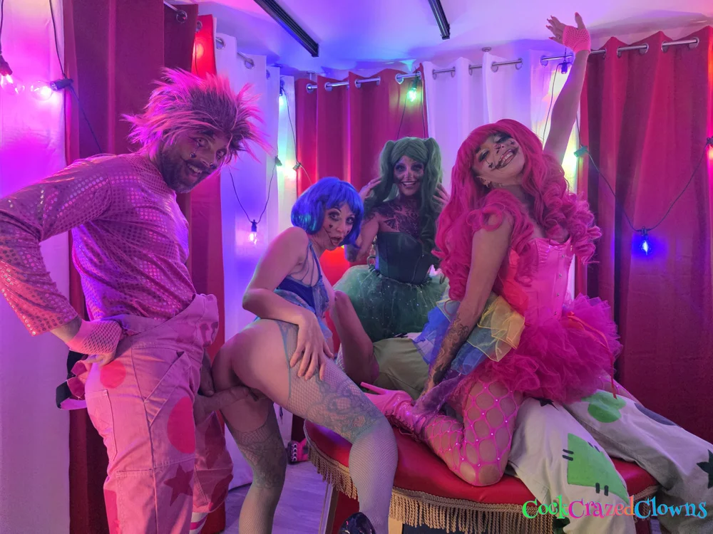 Woman with blue hair bent over a red platform while a man in pink clown overalls stands behind her; two other women in clown costumes pose nearby, one with an arm raised.