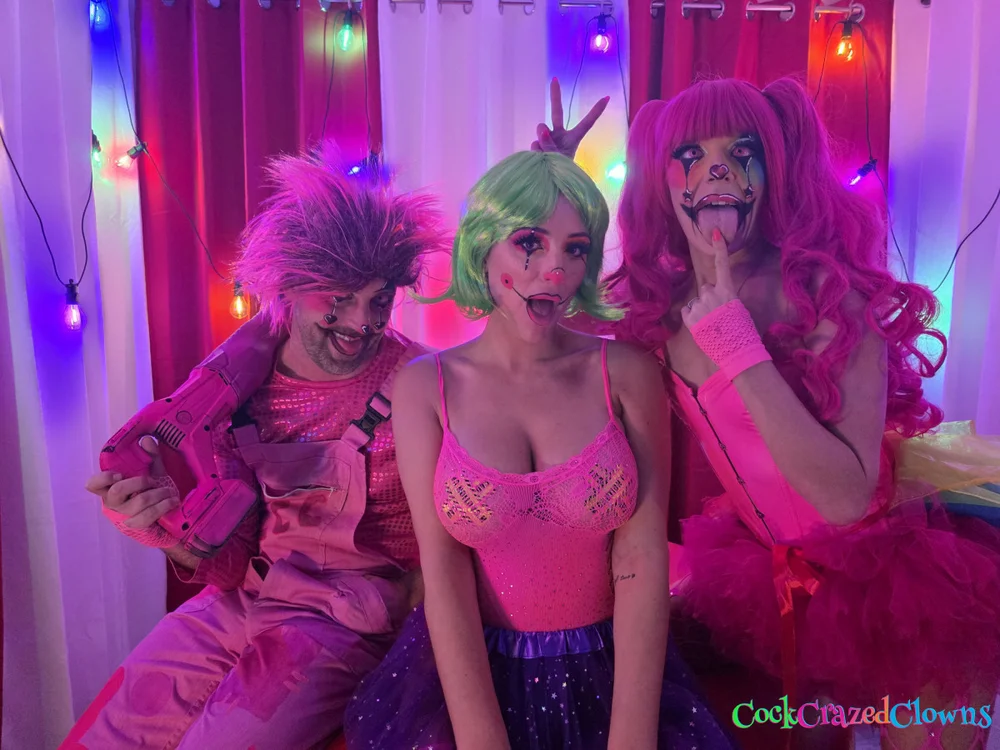 Woman with green bob wig and pink top seated between a man in pink clown overalls holding a pink prop drill and a woman in a pink clown outfit making a face with her tongue out.