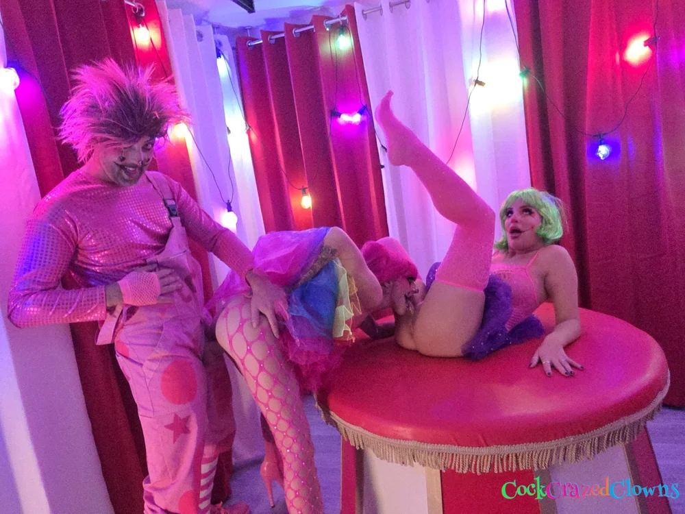 Woman with pink hair bent over a red platform while a woman with green hair performs oral sex on her; a man in pink clown overalls stands nearby watching.