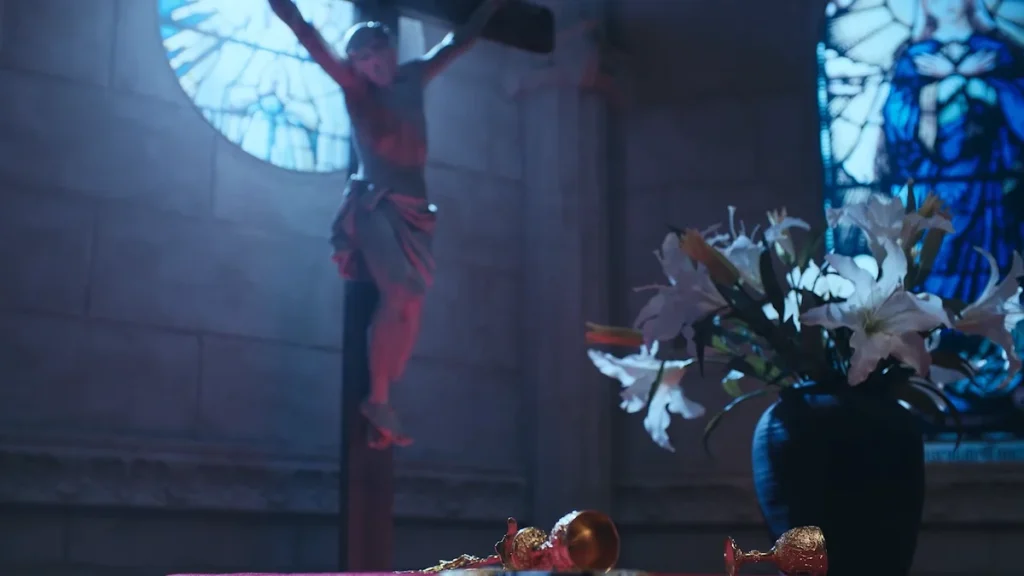 Interior of a dimly lit church with a crucifix statue in the background, stained-glass windows, a vase of white lilies, and golden chalices on a cloth-covered surface in the foreground.