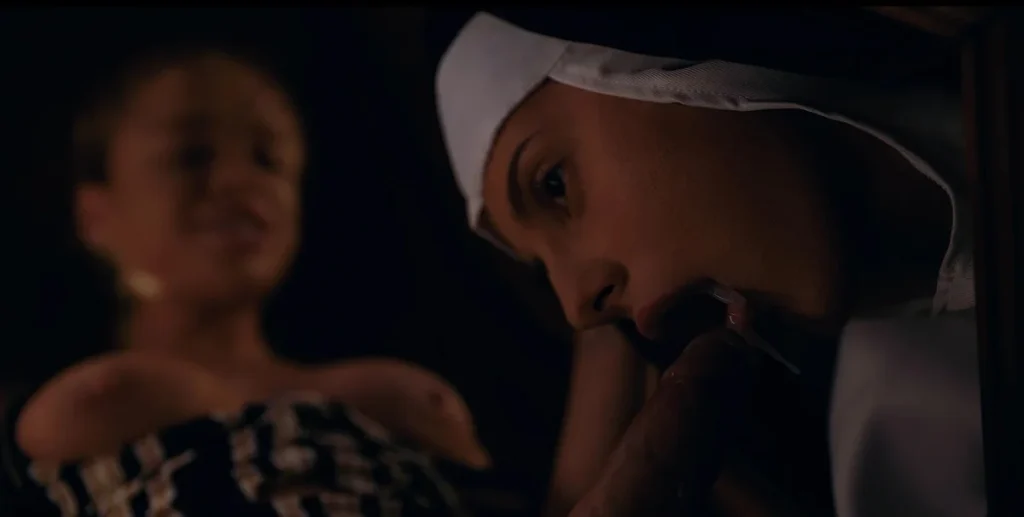 Close-up of a woman in a nun’s habit with her mouth near an erect penis, while a topless woman in a striped top is visible smiling in the background.
