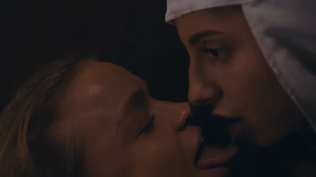 Close-up of two women with their faces close together, tongues extended toward each other, one wearing a white head covering.