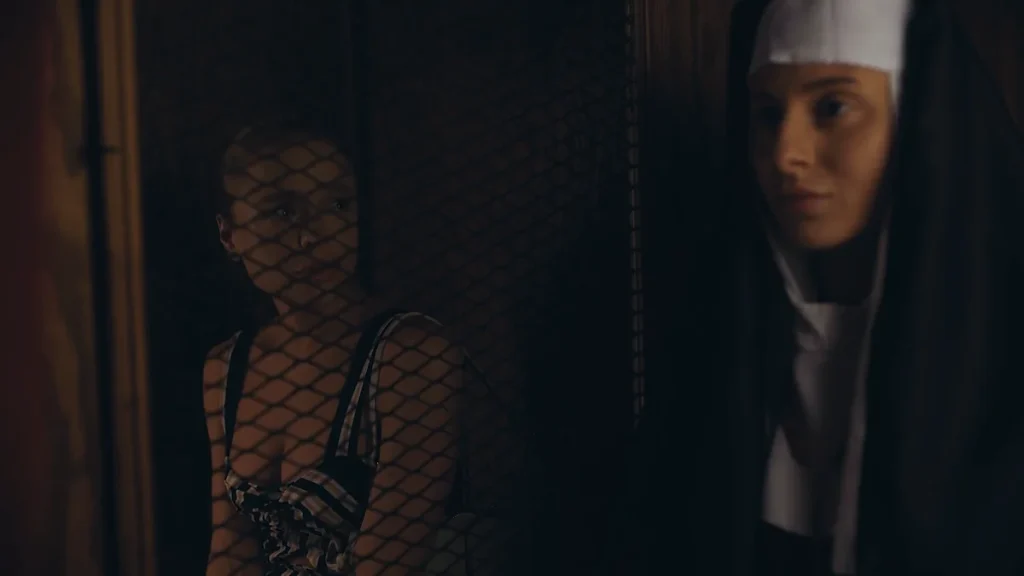 Woman with blonde hair in a striped top looking through a wire mesh from inside a dark enclosure, while a woman in a nun’s habit stands outside looking toward her.