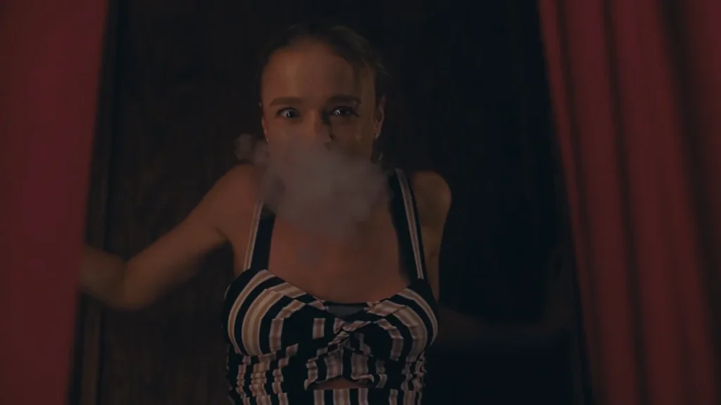 Woman with blonde hair in a striped crop top exhaling a cloud of smoke while looking at the camera in a dark room with red curtains.
