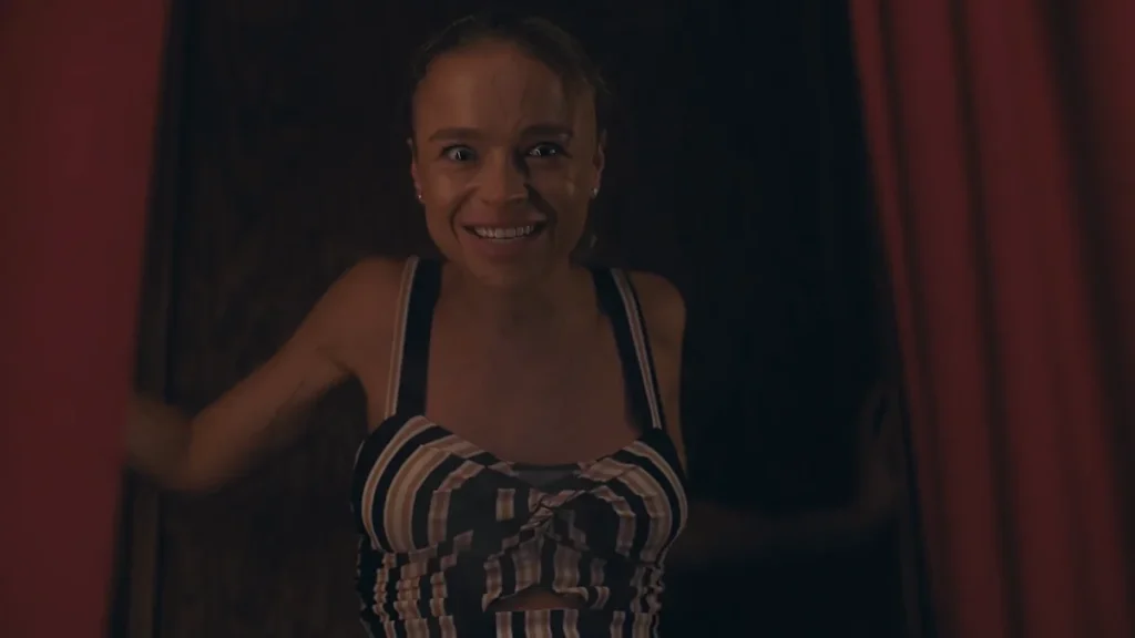 Woman with blonde hair in a striped crop top smiling widely with an intense expression while looking at the camera in a dark room with red curtains.