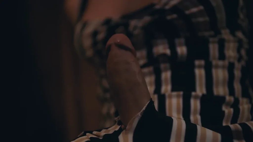 Close-up of an erect penis in the foreground with a woman in a striped top partially visible in the background.