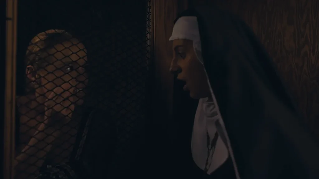 Woman with blonde hair looking through a wire mesh while a woman in a nun’s habit stands close on the other side, facing her in profile.