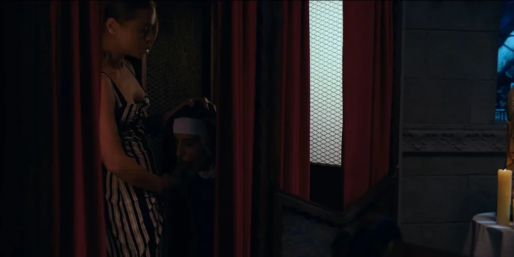 Woman in a striped dress standing while a woman in a nun’s habit kneels in front of her performing oral sex, in a dimly lit area with red curtains and a candle visible.