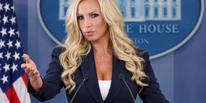 Blonde woman as white house press secretary stand at the podium with arm raised.