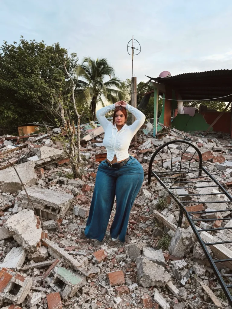Woman with reddish-brown hair standing amid rubble and debris, wearing a tight white long-sleeve top and blue wide-leg jeans with a black belt, hands clasped behind her head, looking at the camera; damaged structures, metal bed frame, and trees visible in the background.