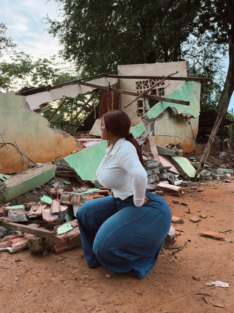 Woman with reddish-brown hair in a ponytail kneeling on dirt ground in front of collapsed building walls, wearing a tight white long-sleeve top and blue wide-leg jeans, body turned sideways with one hand on her hip, looking to the side.