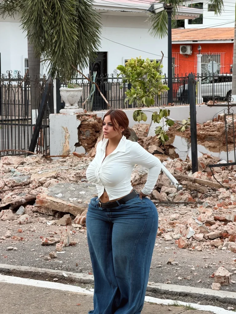 Woman with reddish-brown hair standing on a street amid rubble, wearing a tight white long-sleeve top and blue wide-leg jeans with a black belt, one hand on her hip, looking at the camera; damaged wall, fence, and houses visible behind her.