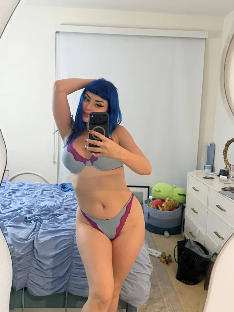 Woman with blue hair taking a mirror selfie in a bedroom, wearing a light blue bra and matching panties with pink lace trim, one arm raised behind her head.